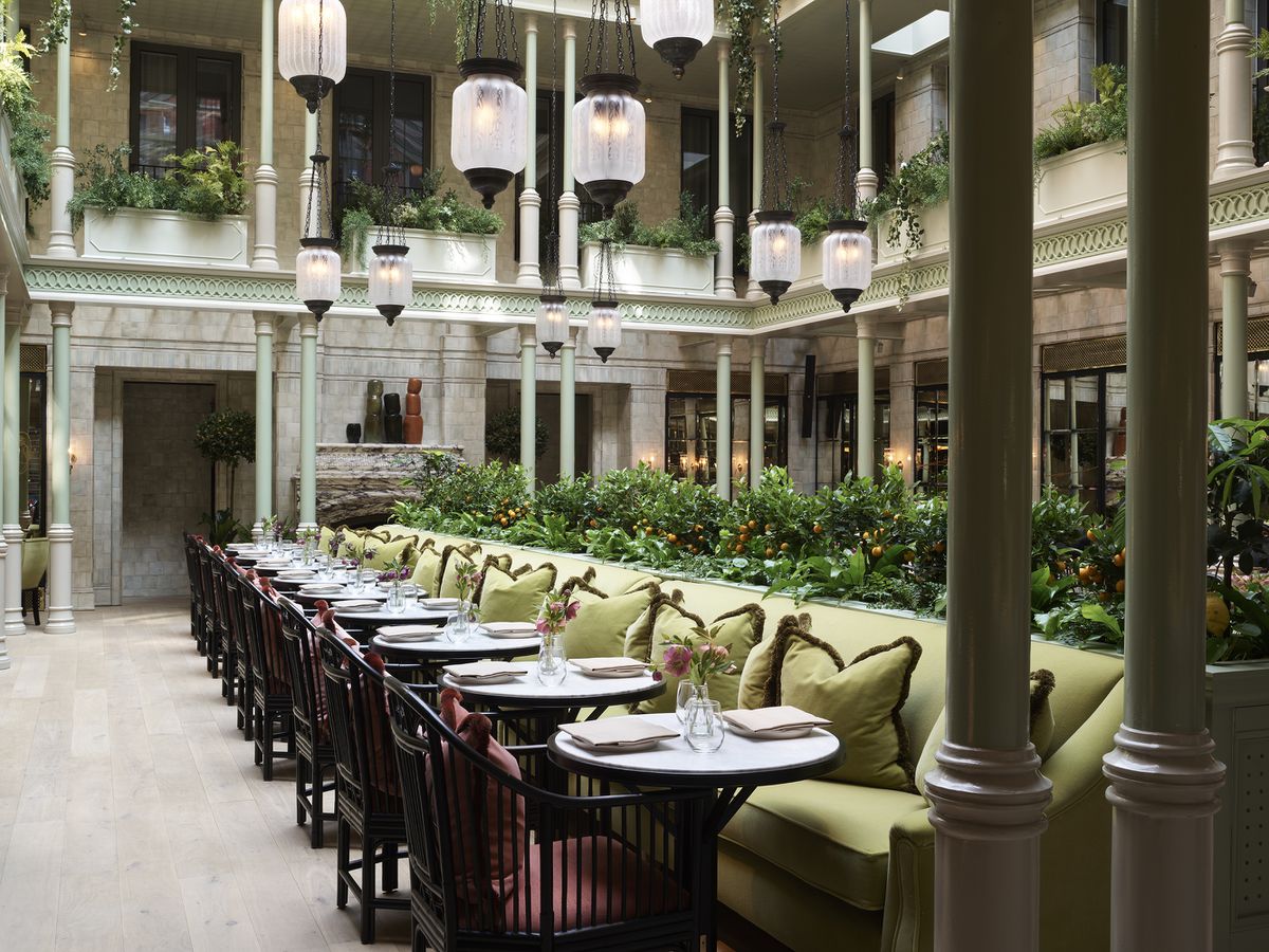  In a plant-filled restaurant dining room inside an atrium, there’s a row of small round tables with a single, long green banquette on one side and chairs on the other.
