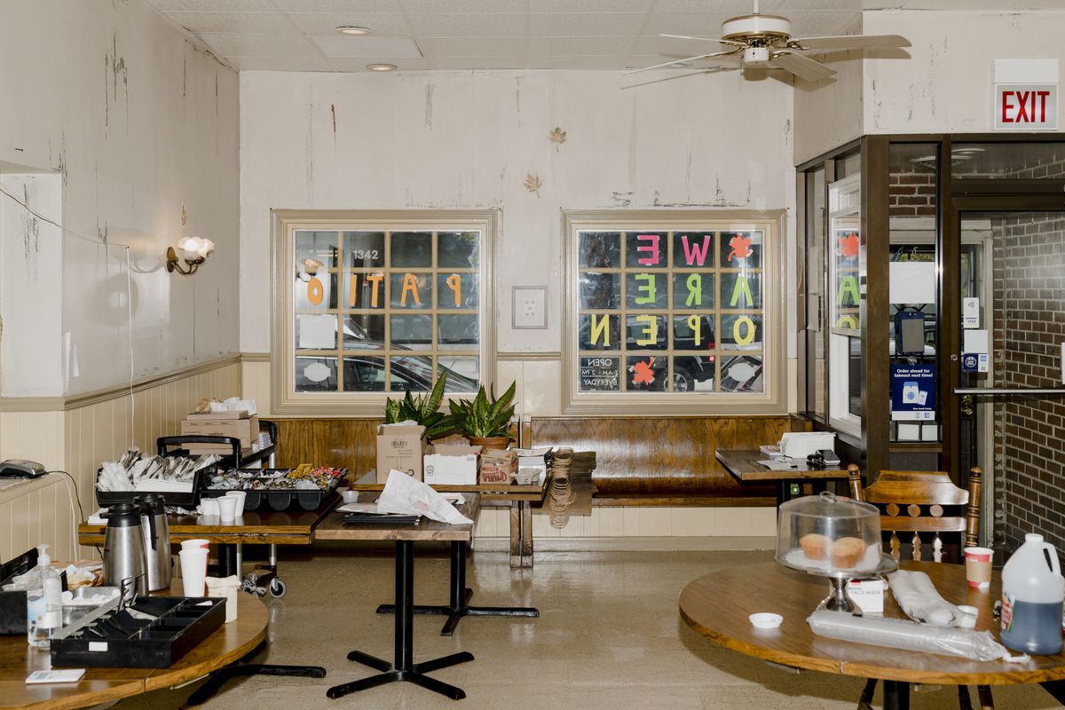  A sparsely decorated room with beige walls with two street-facing windows with grid panes. The room is filled with tables placed with packaged plastic cutlery, condiments, coffee carafes, and muffins in a cake stand. The vibe is understated but welcoming.