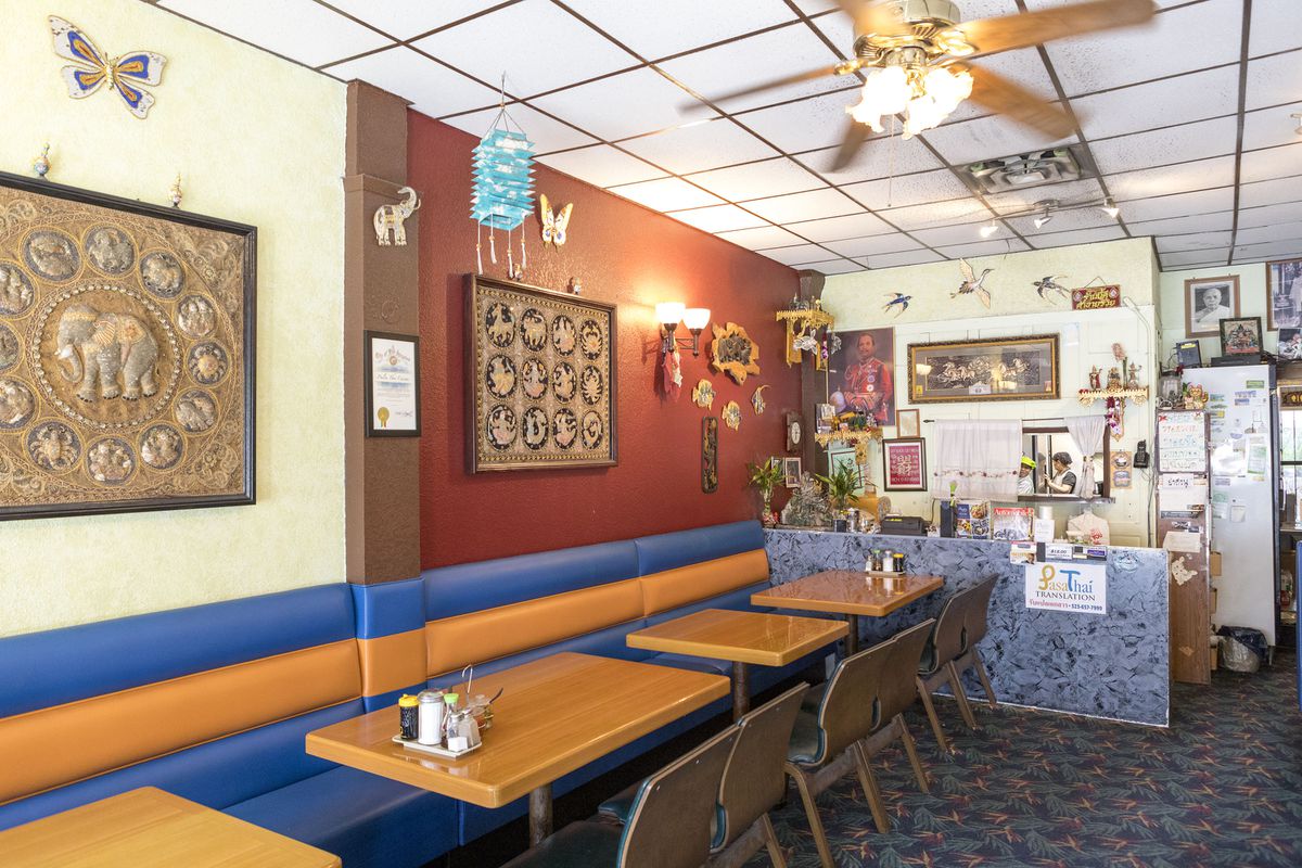  A side view of the inside of a restaurant facing a register stand. There are half booths wrapped in vinyl, with wooden tables and chairs. The walls are decorated with mismatched antique looking art featuring elephant motifs.